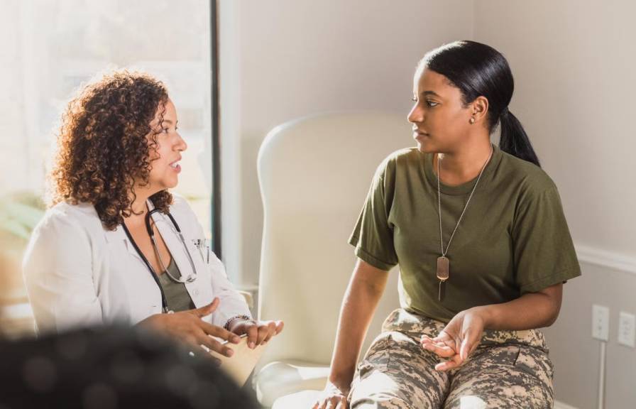 A woman in a medical coat speaks to a soldier in a green shirt and camo pants