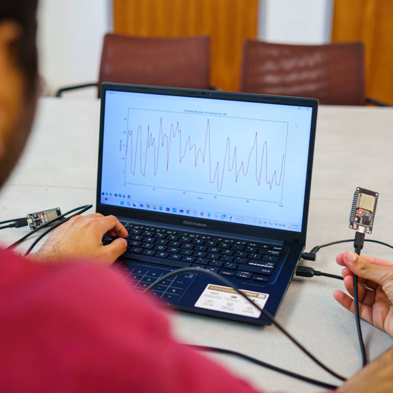 A researcher holds a wifi device in front of a laptop that displays heart rate information.