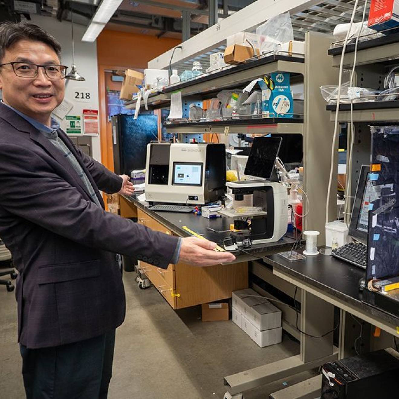 Man in a dark gray suit stands in a lab in front of multiple machines on a benchtop