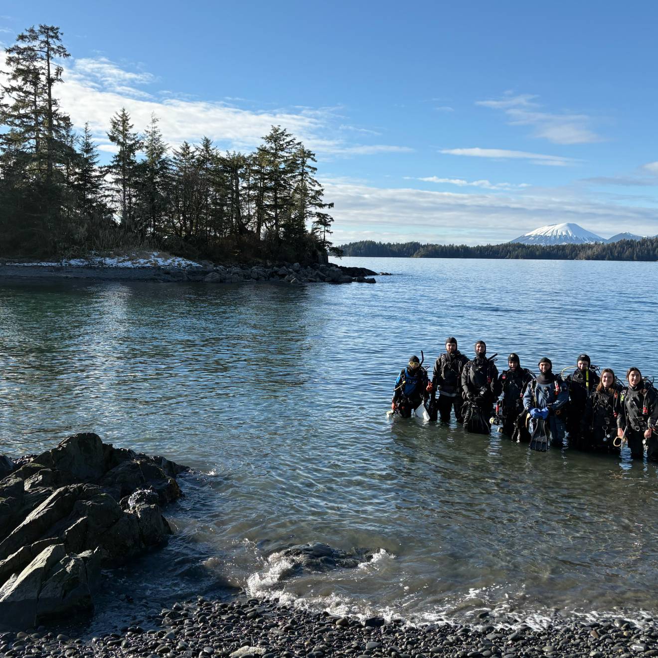 A smiling group of people stands in the water in wetsuits and SCUBA gear, with evergreen trees and a snow-covered peak in the background