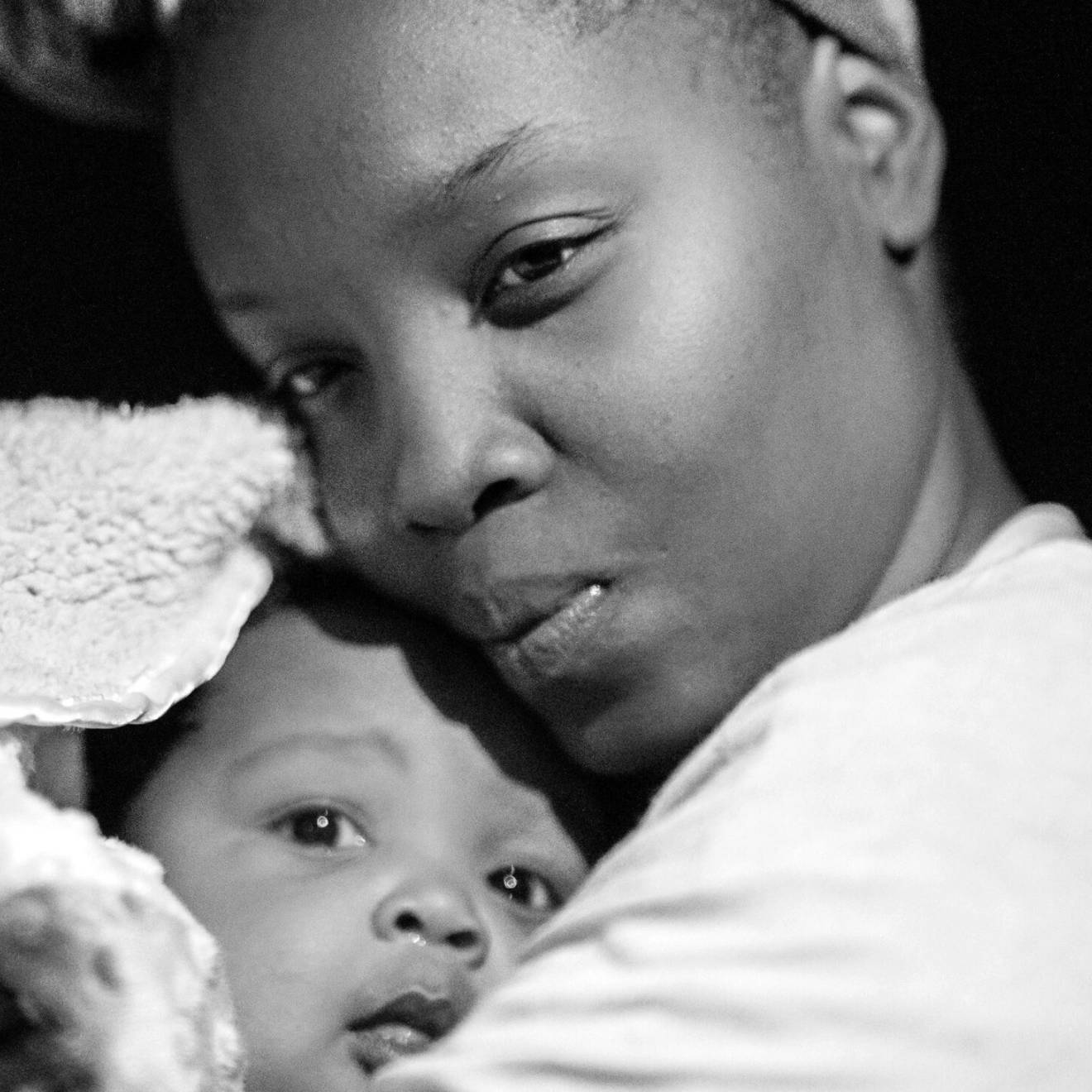 In a black-and-white close-up photo, a Black woman gazes thoughtfully as she coddles a baby wrapped in a blanket