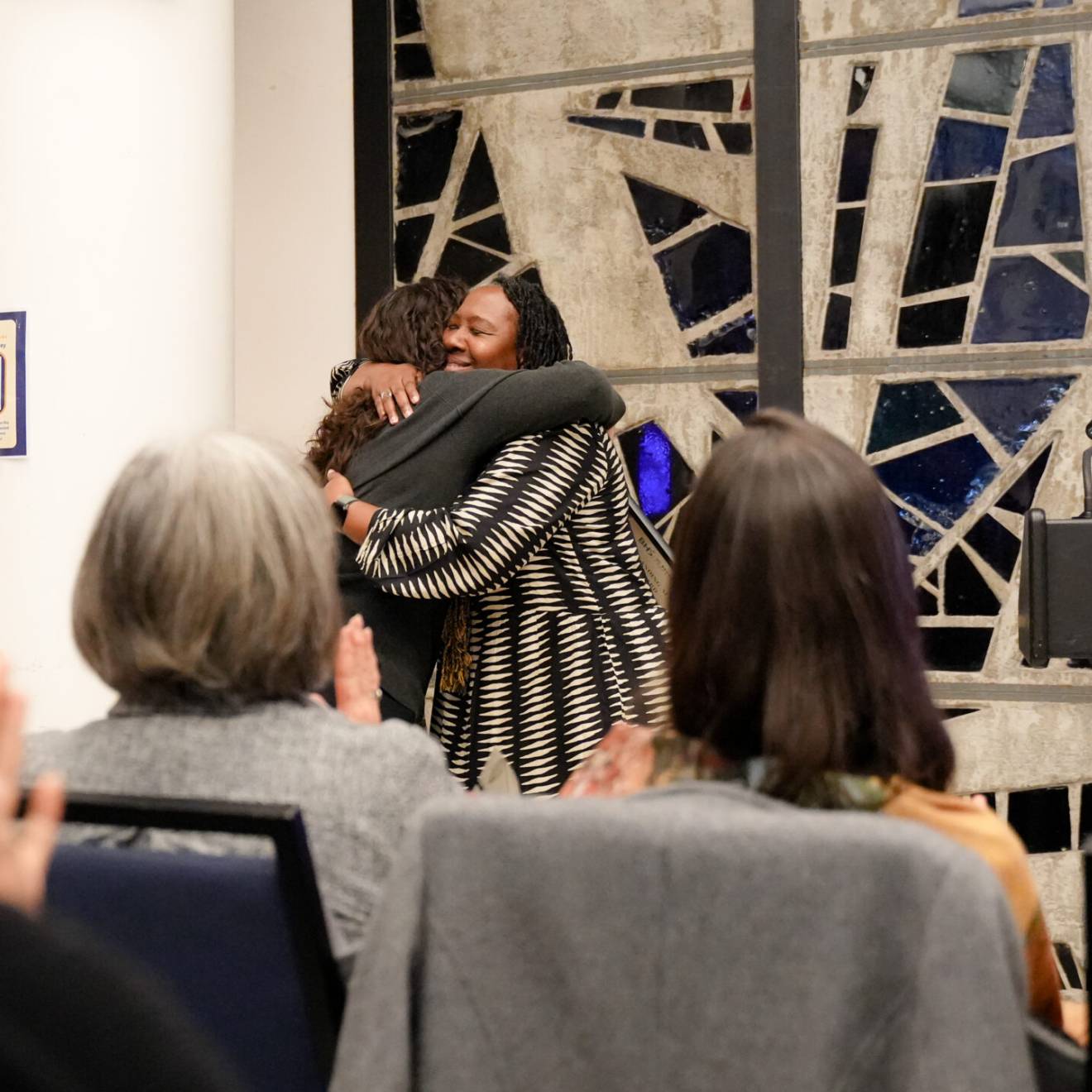 two women hug in front of a stained glass window as others applaud