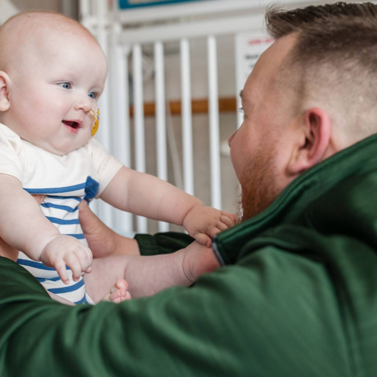 A man in a green jacket looks at a baby in front of him, both looking excitedly at each other