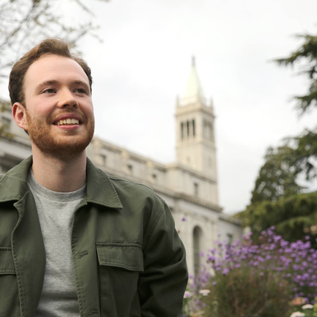 a young man wearing a green jacket puts his hands in his pockets and smiles in front of the Campanile