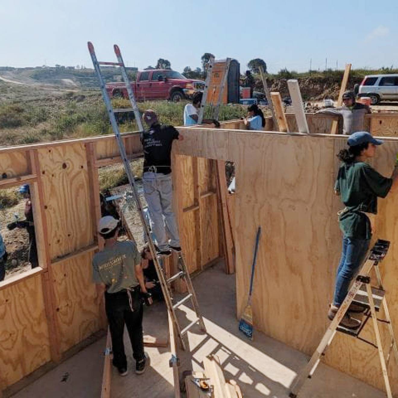 workers, some on ladders, building a house in an unpopulated, scrubby landscape