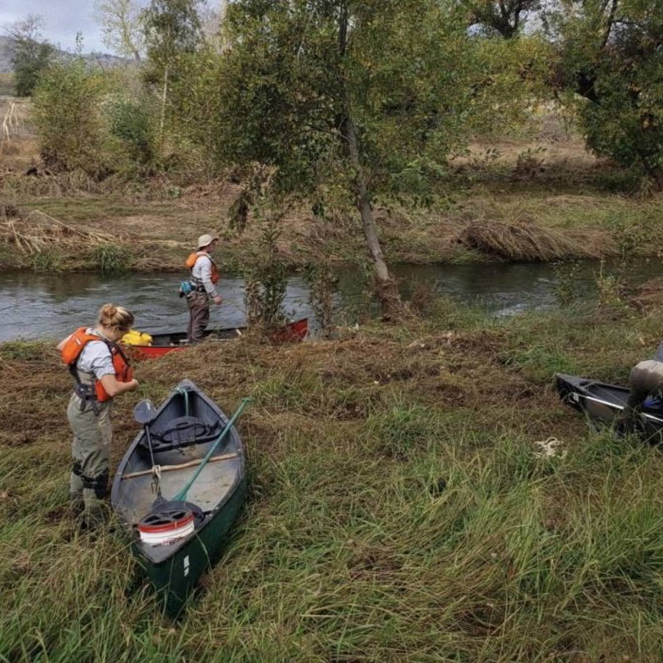 Three people with water gear and canoes on the bank of a creek
