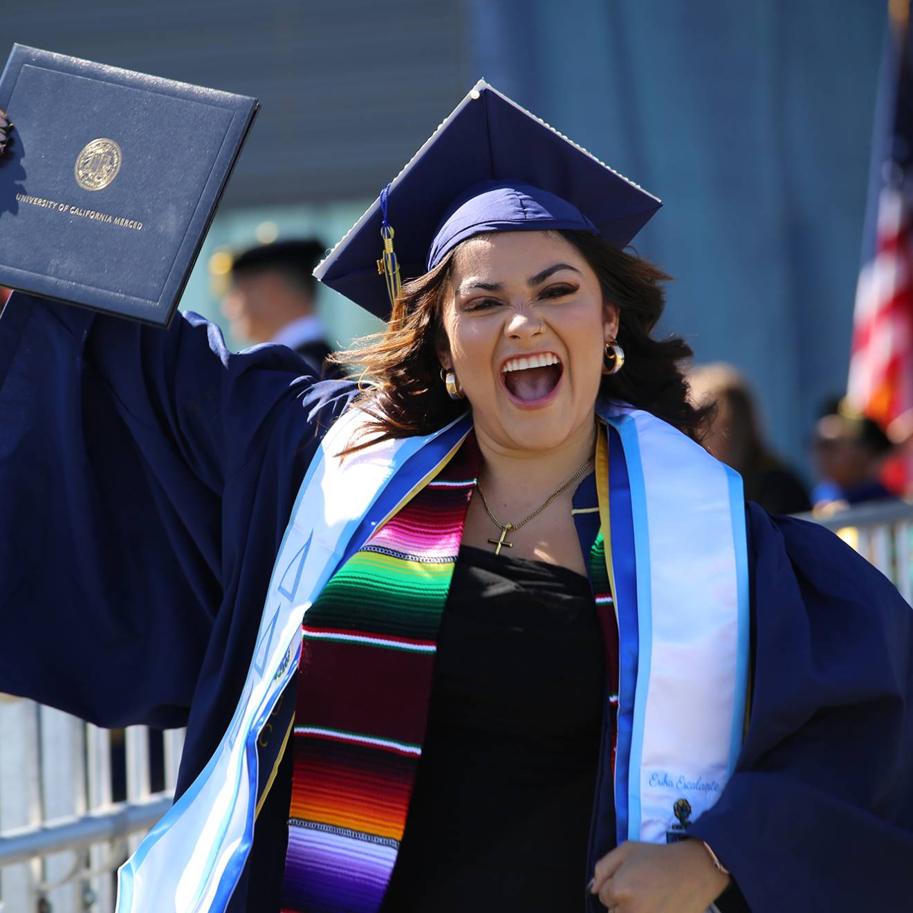 A woman in a cap and gown smiles while holding up her diploma