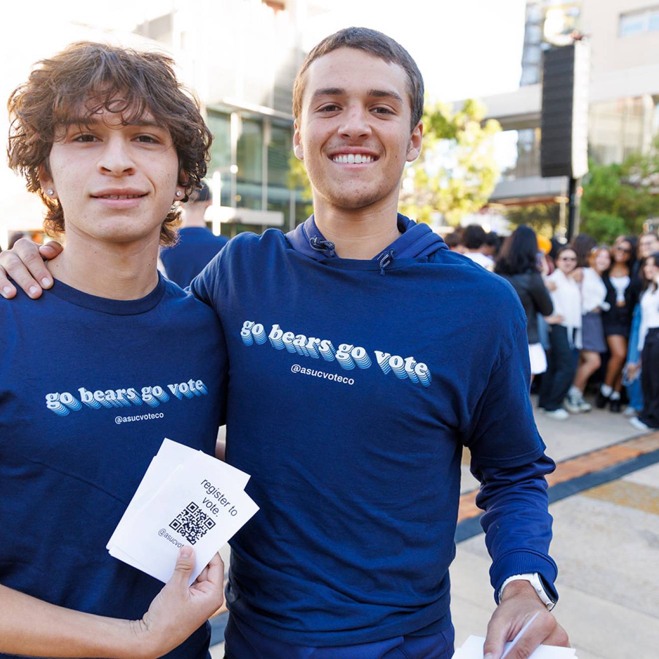 Two students smile for the camera with arms around each other's shoulders, wearing matching t-shirts that read "Go Bears Go Vote." One student holds a stack of printed QR codes labeled "Register to Vote"