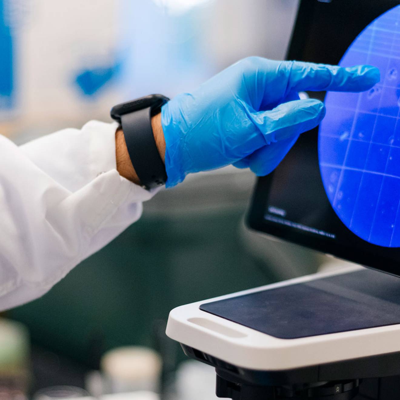 A hand in a blue glove points to a tablet screen showing a microscope view.