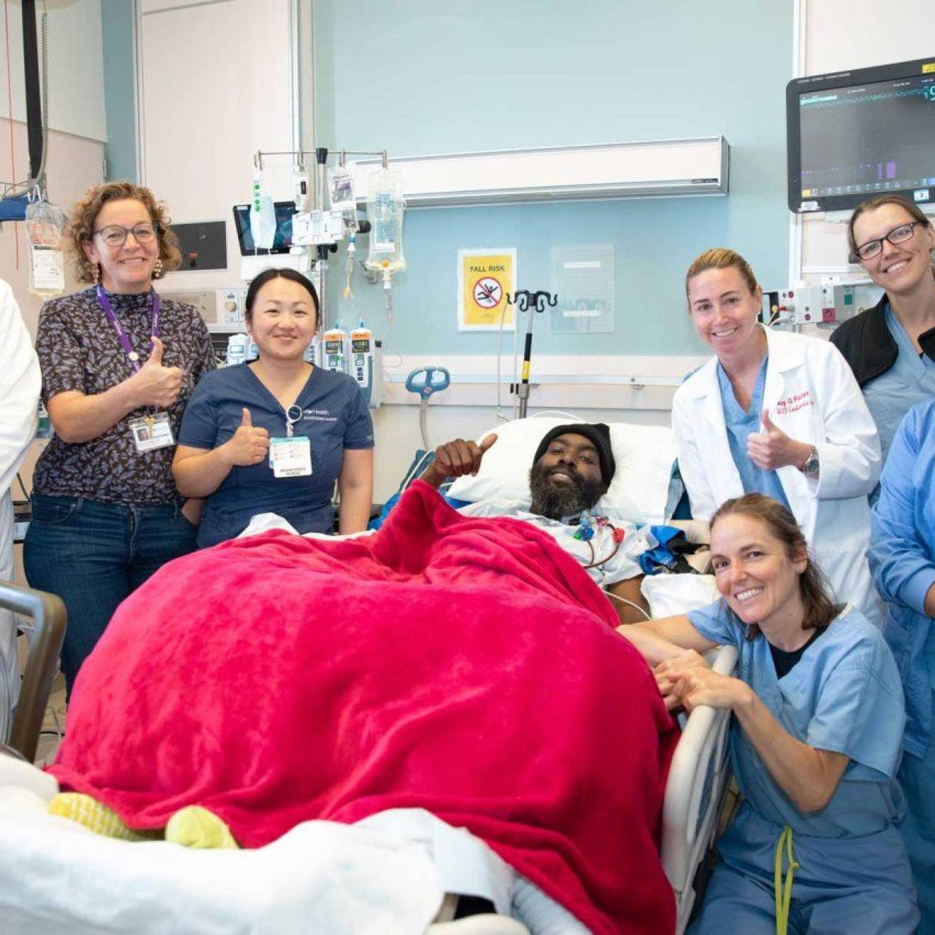 A man in a hospital bed with a red blanket on him is surrounded by a team of nurses and doctors, smiling and giving thumbs up