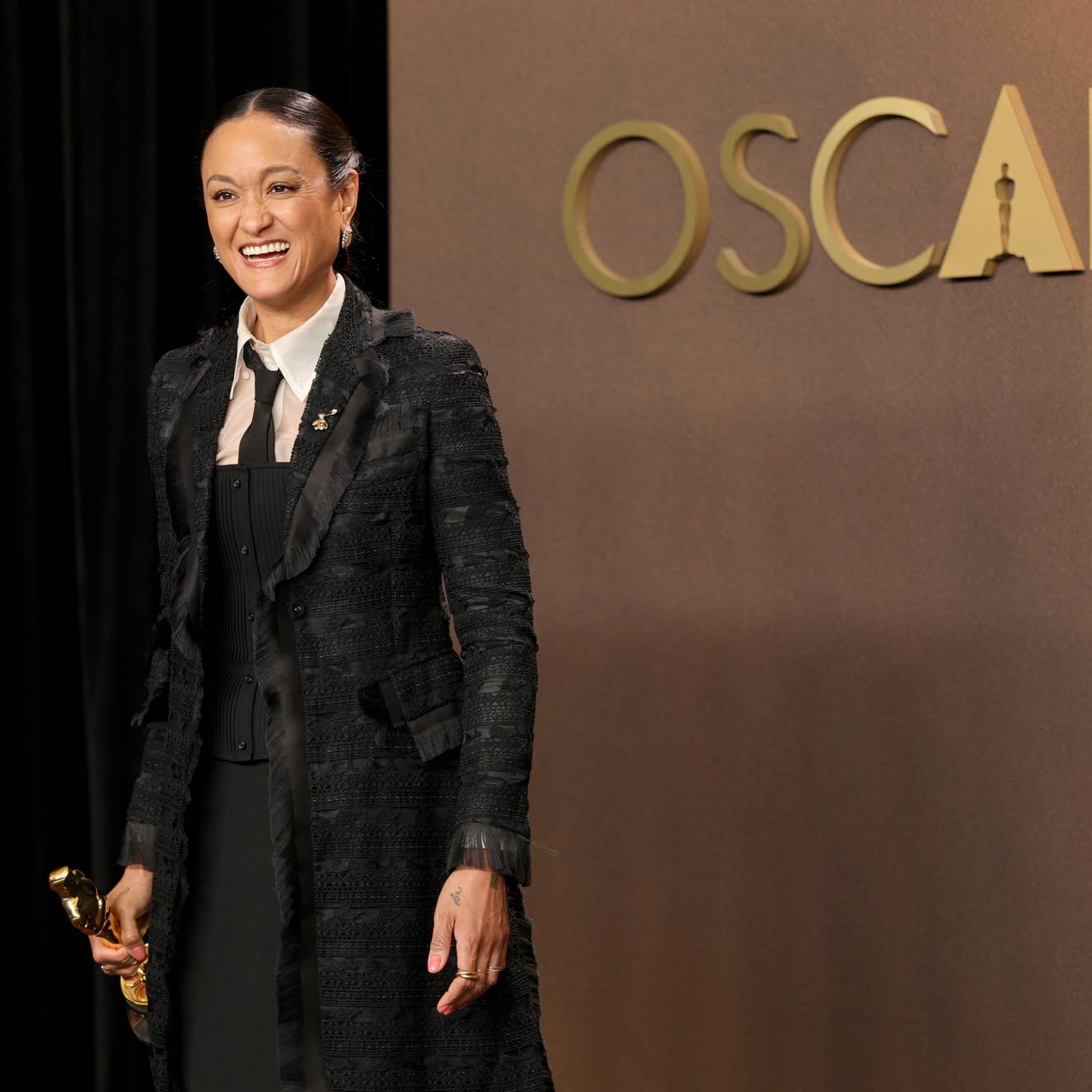 A woman in a black coat holds an Oscar smiling overjoyed in front of a gold background that says Oscars