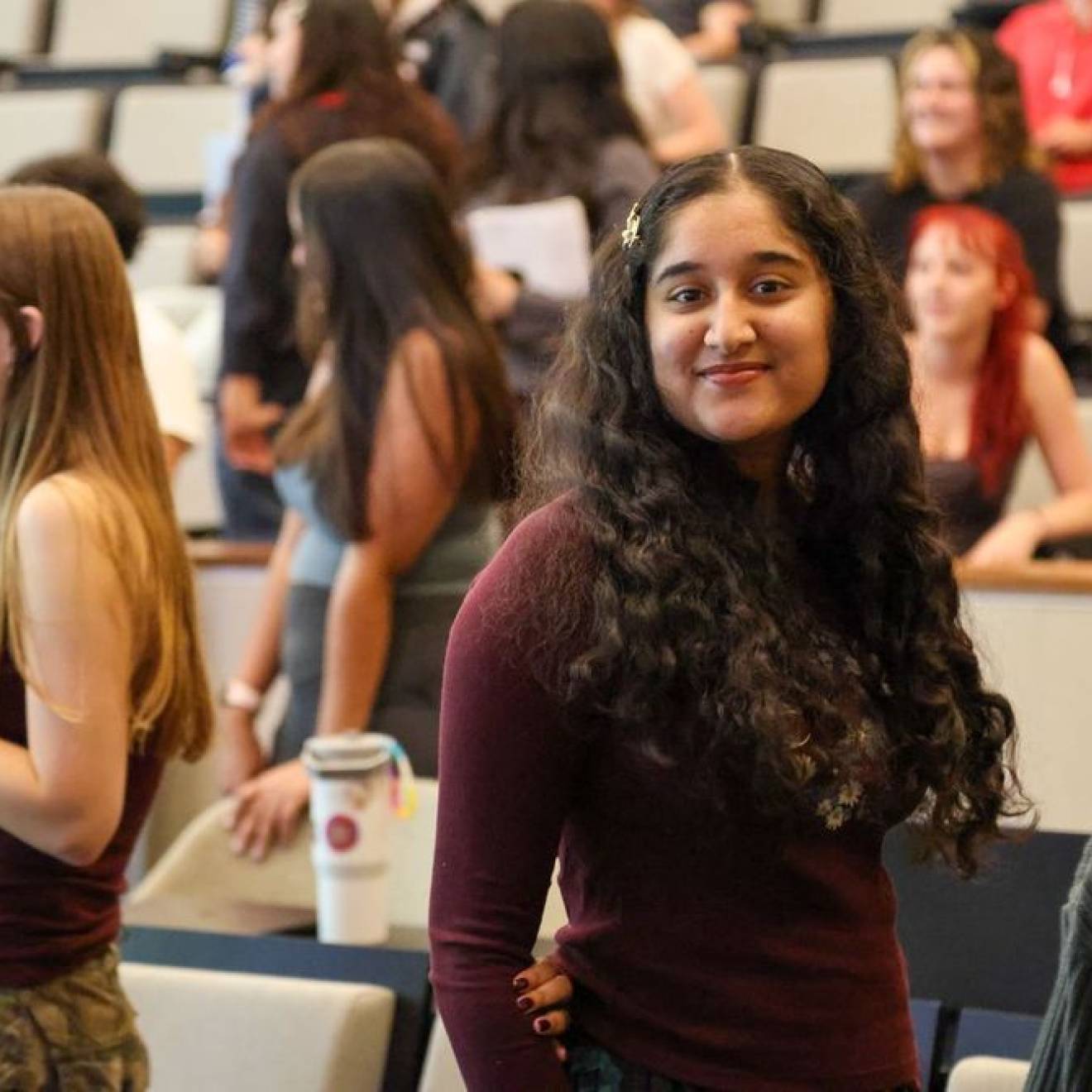 A young woman smiles at the camera with other students standing smiling looking away from the camera behind her