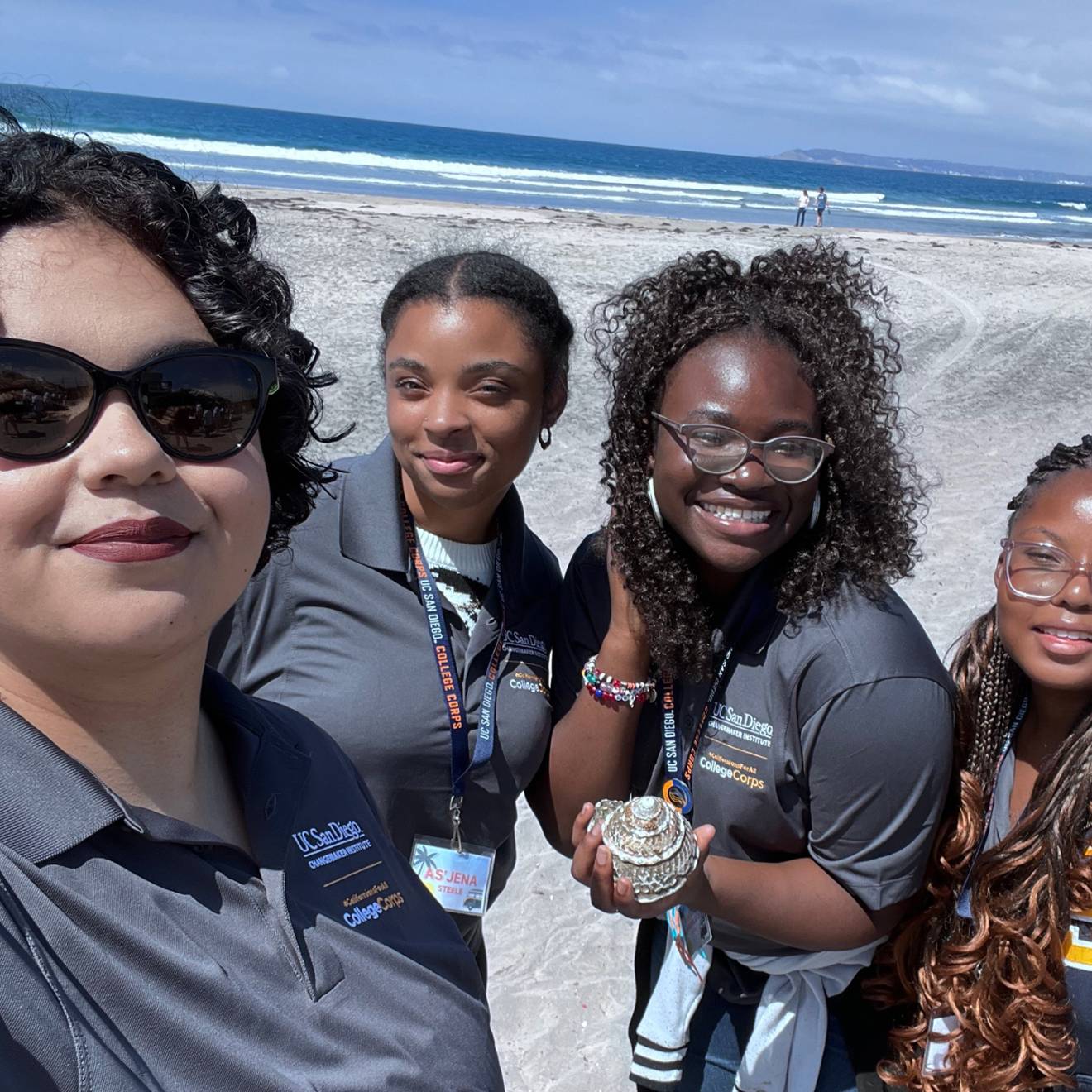 Four women in gray UC San Diego College Corps polo shirts take a selfie on the beach, smiling