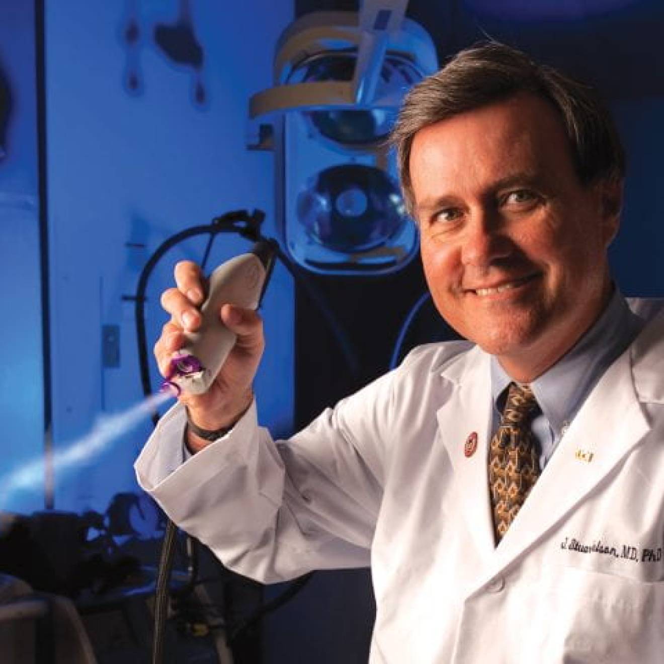 A surgeon in a white coat in a darkened lab holds a skin cooling device up to the camera and smiles.