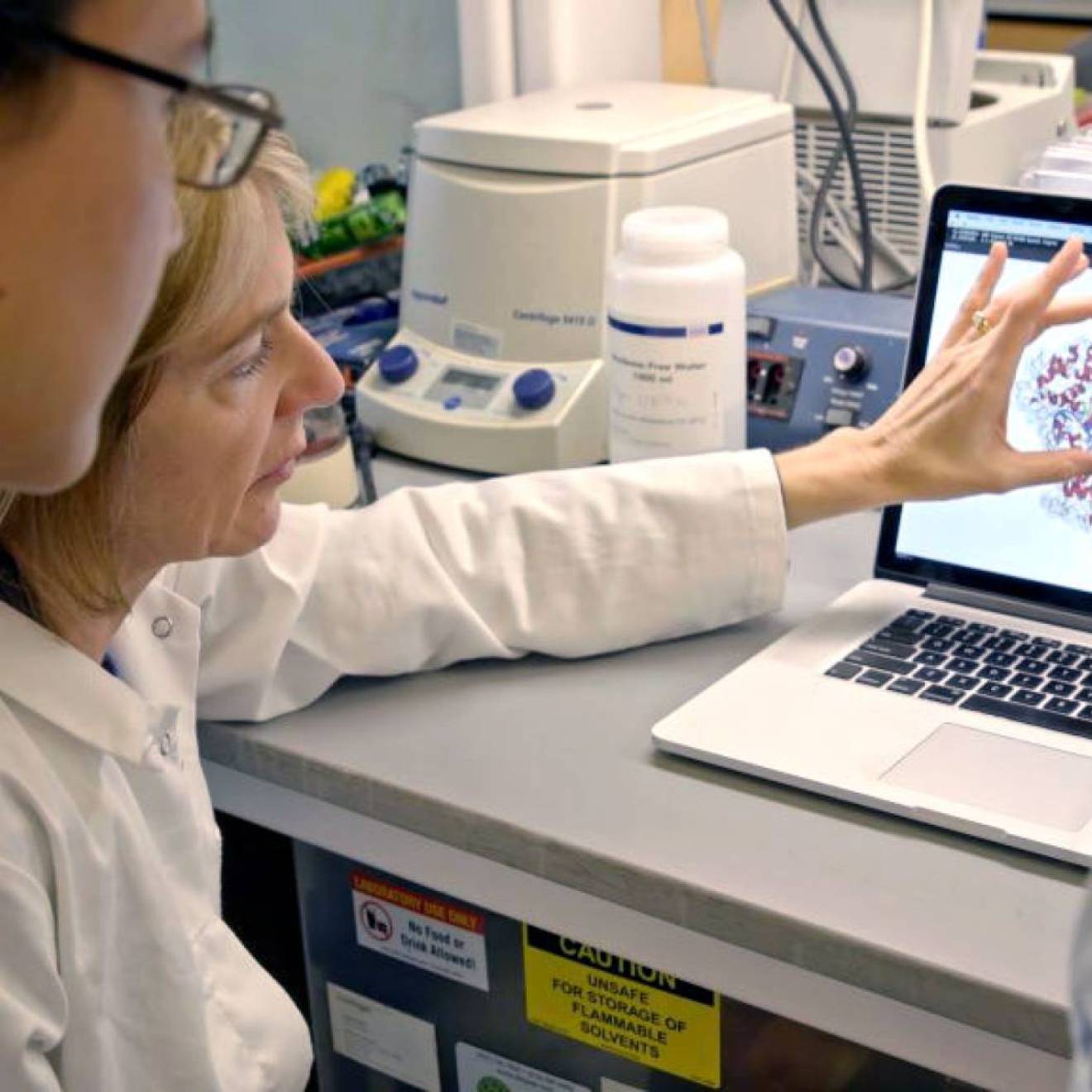 A woman doctor in a lab coat shows a male student a molecule on a computer