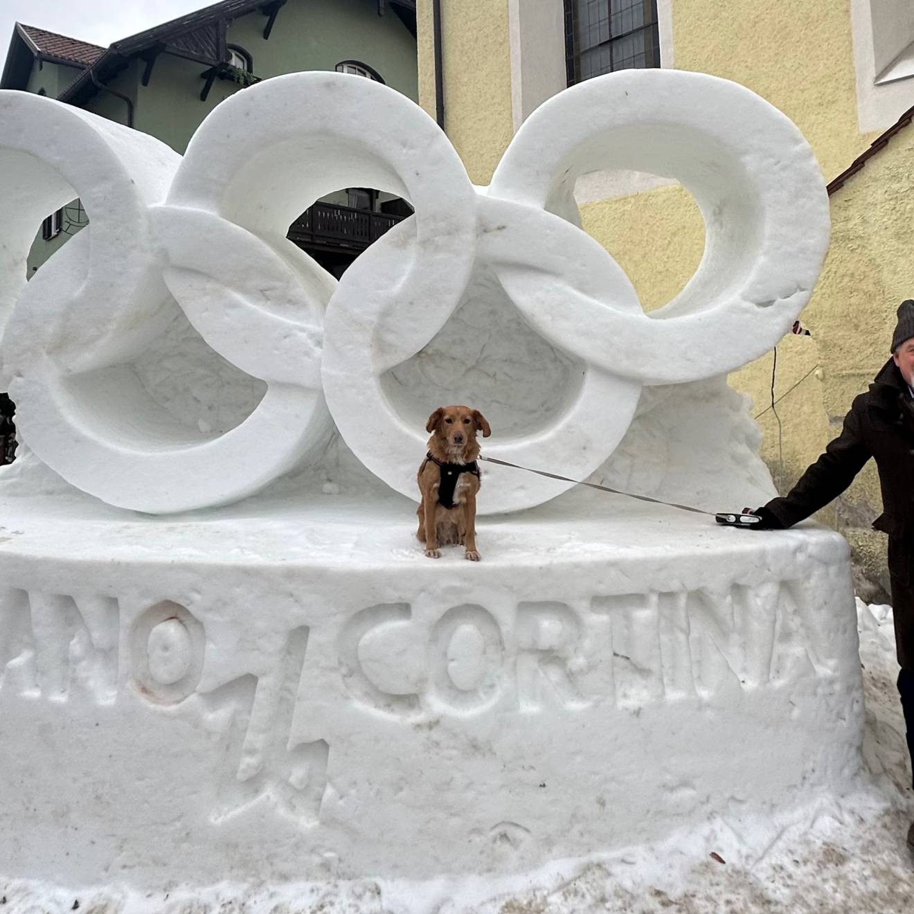 In front of a snow sculpture of the Olympic rings that reads Milano Cortina, a man stands next to a service dog sitting on the sculpture