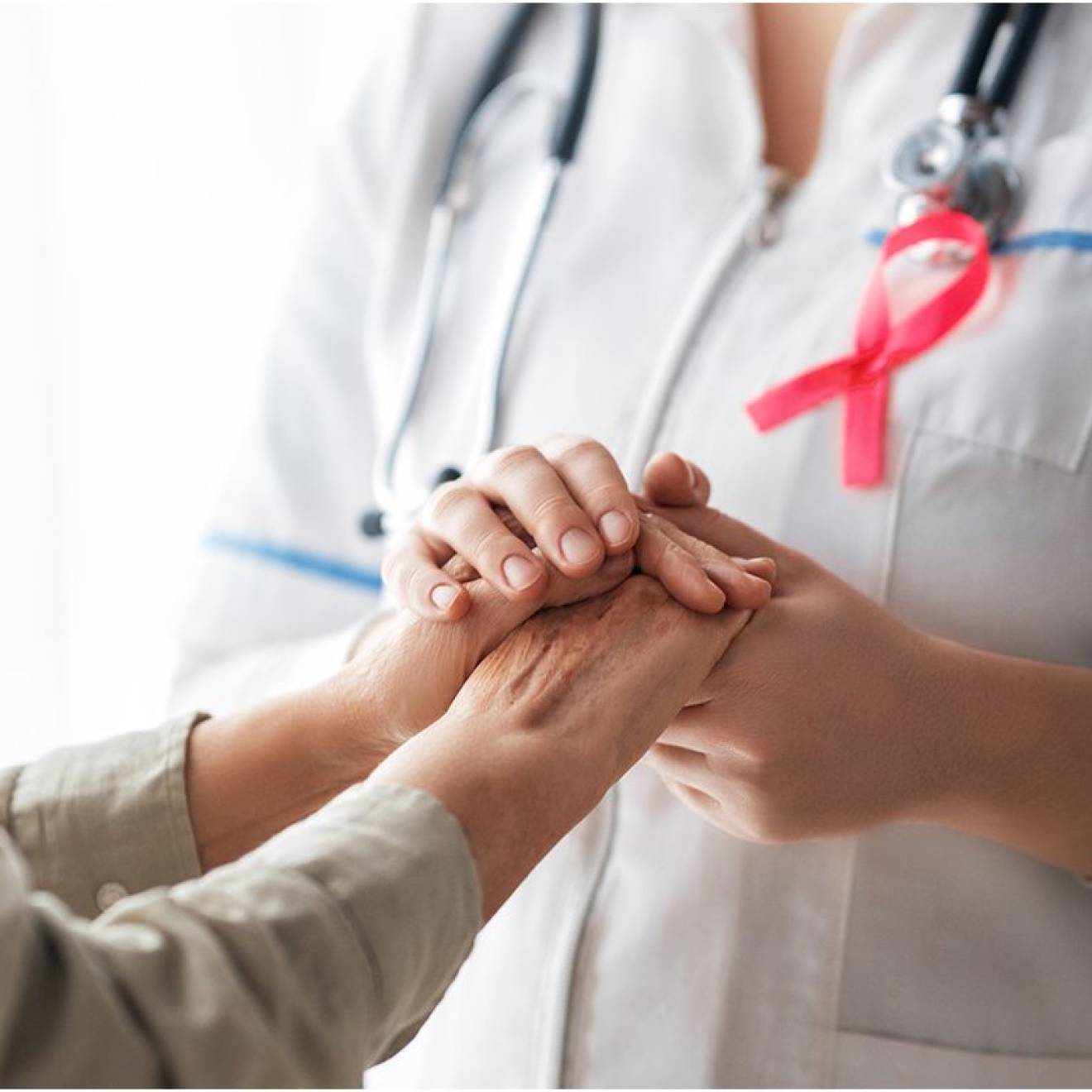 A patient, left, and medical care provider holding hands; provider has a pink breast cancer ribbon on her lapel