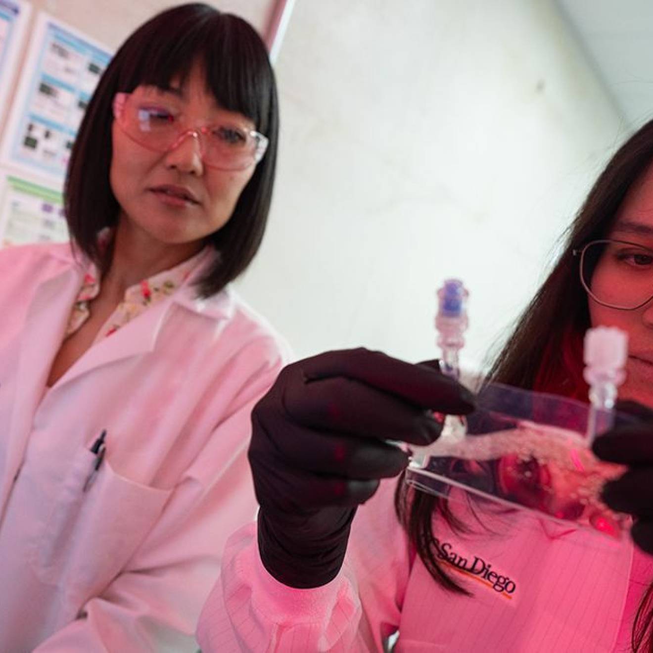 Two women in lab coats in a lab with reddish lighting in the corner; one holds and looks at a small baggie