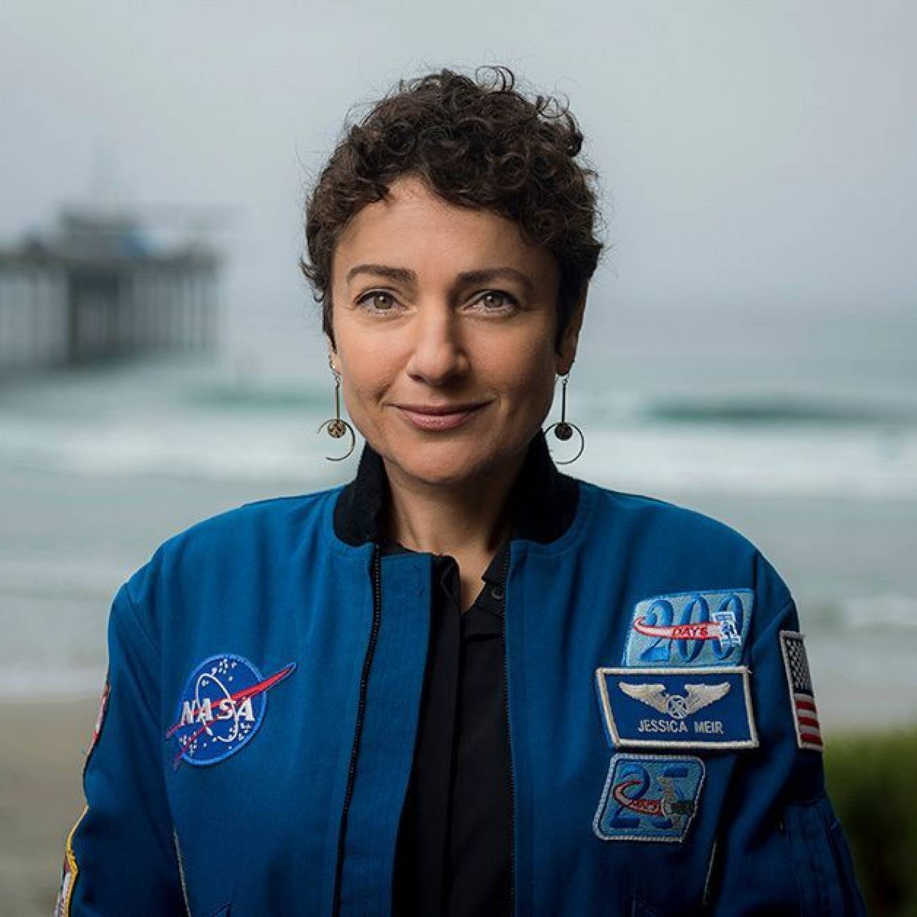 A woman in a blue NASA jumpsuit photographed with a pier and ocean behind
