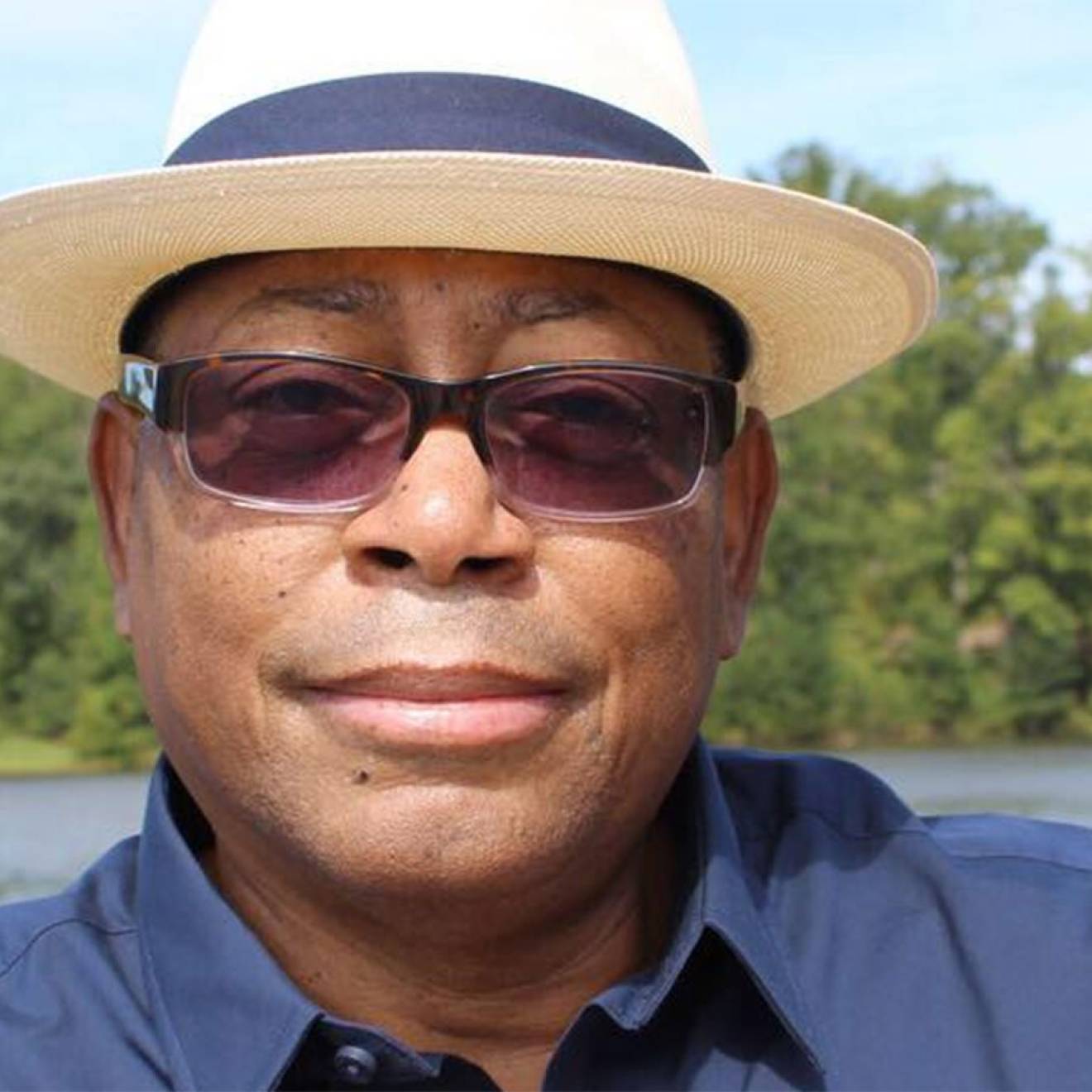 Mel Mann, wearing sunglasses and a fedora, smiles faintly, standing on the shore of a lake