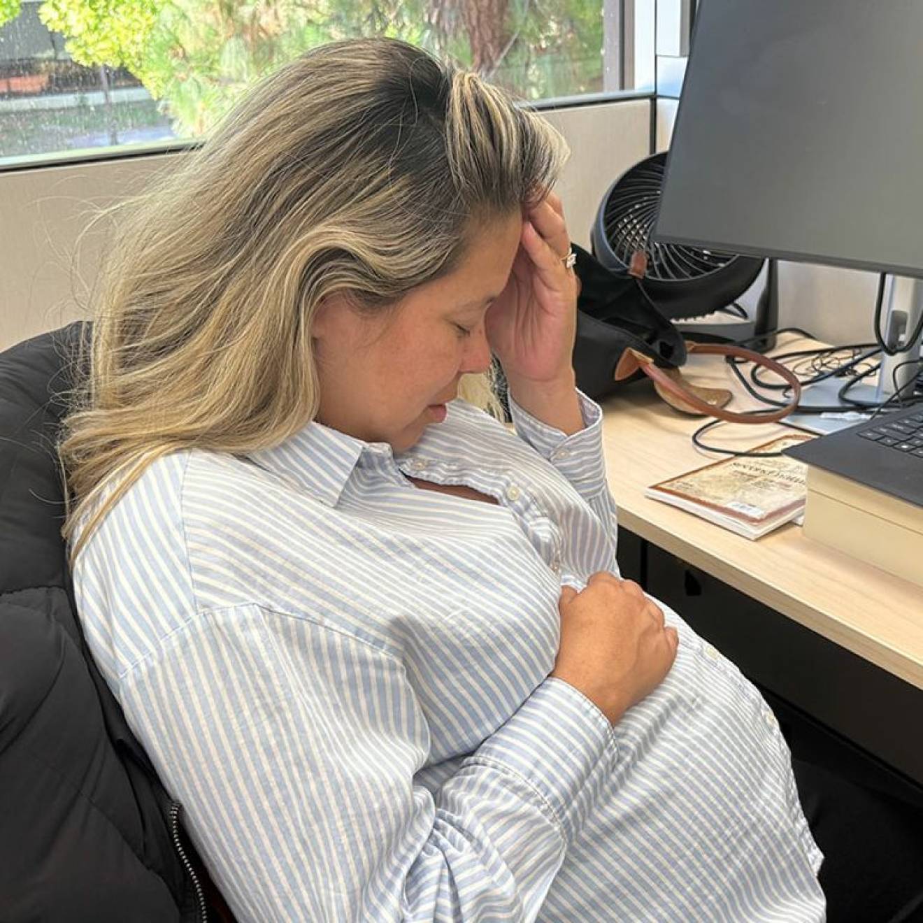 A woman holds her head with another hand over her pregnant belly while sitting at a desk in an office
