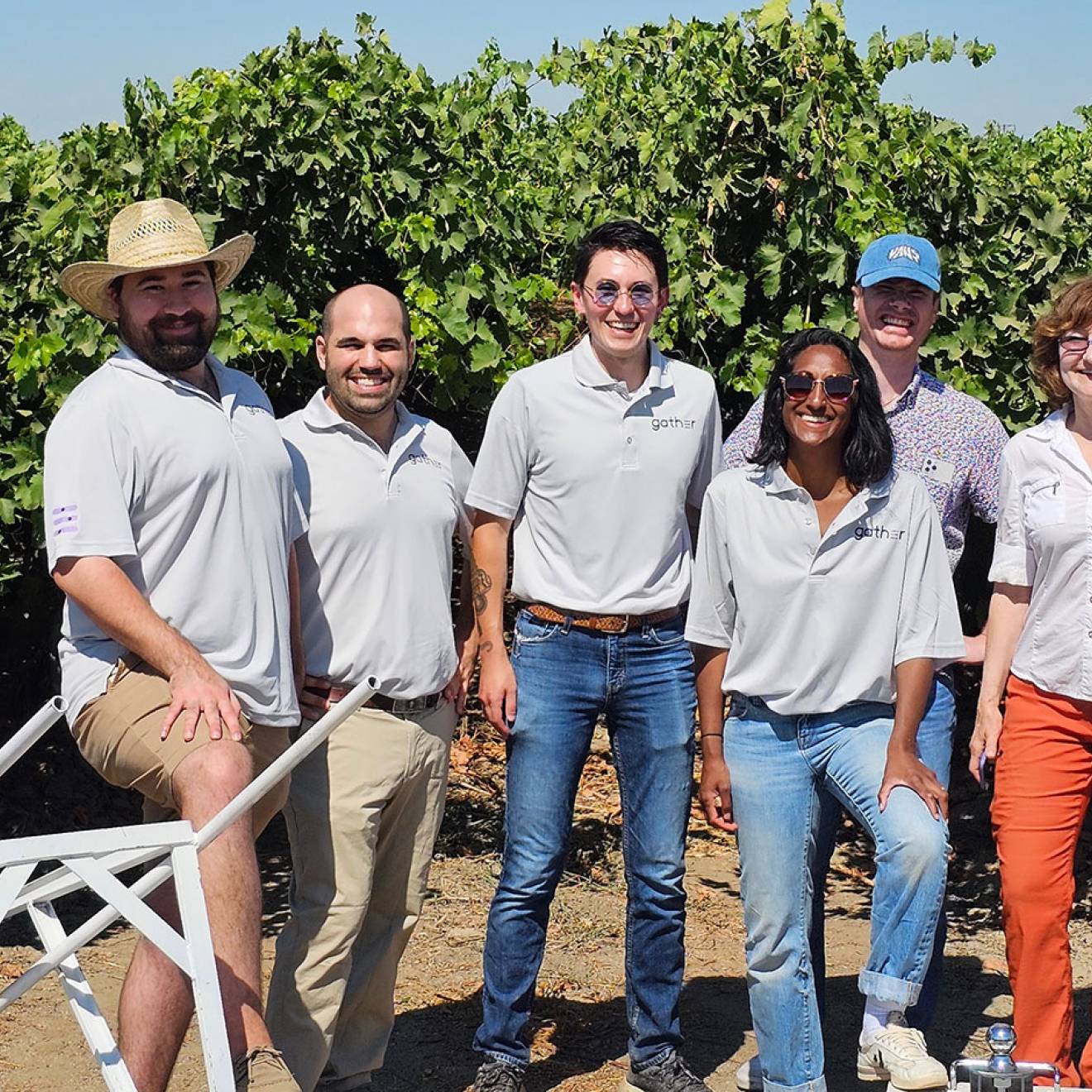 10 employees of Milano Technical Group smile for an informal photo, many wearing matching gray polo shirts, standing in a Central Valley orchard, behind a few pieces of machinery designed by the company. 
