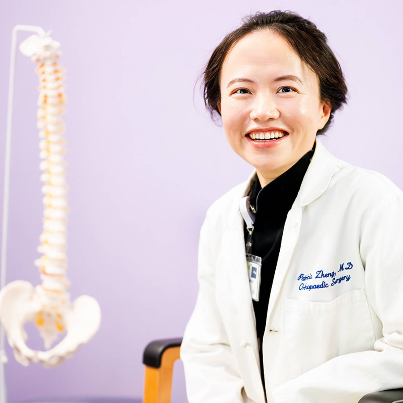 A woman with short hair in a white doctor's coat smiles in a chair with a model of a human spine behind her against a lilac backdrop