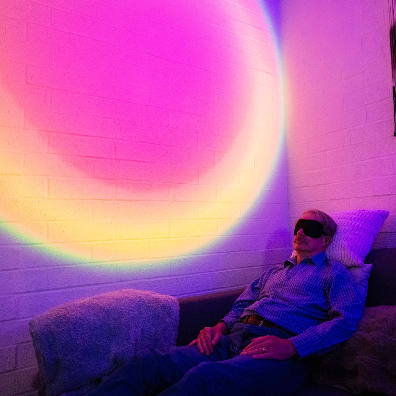 A man wearing an eye mask reclines on a couch in a darkened room. Behind him, the wall is illuminated with a circular, rainbow-colored light