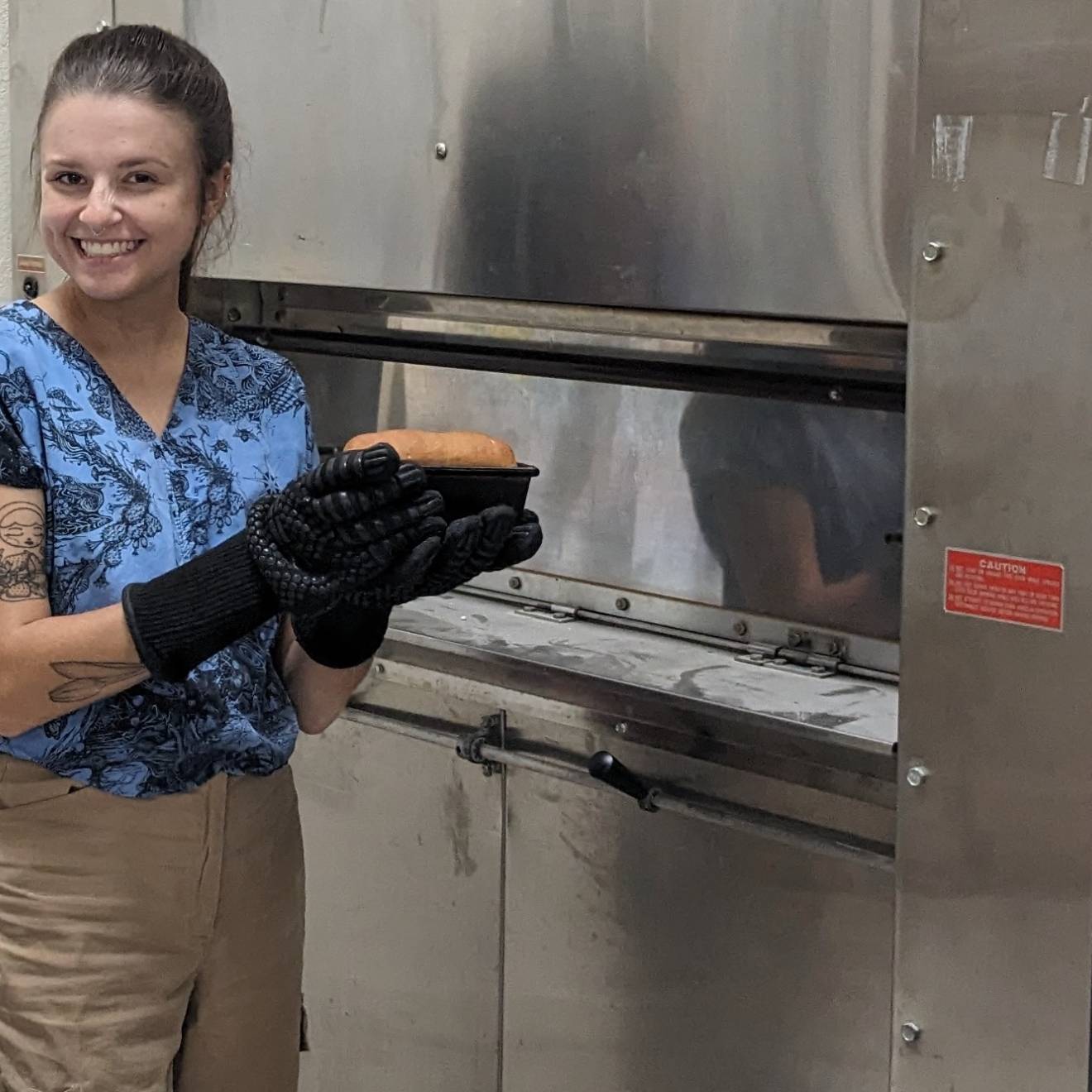 Woman with baking mitts smiles and holds up a loaf of bread in front of an oven