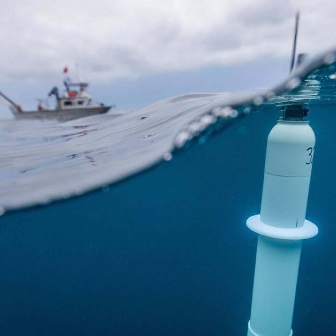 Taken with a lens that's half submerged, a photo of a cylindrical instrument floating near the surface of the ocean in the foreground, with a ship in the background