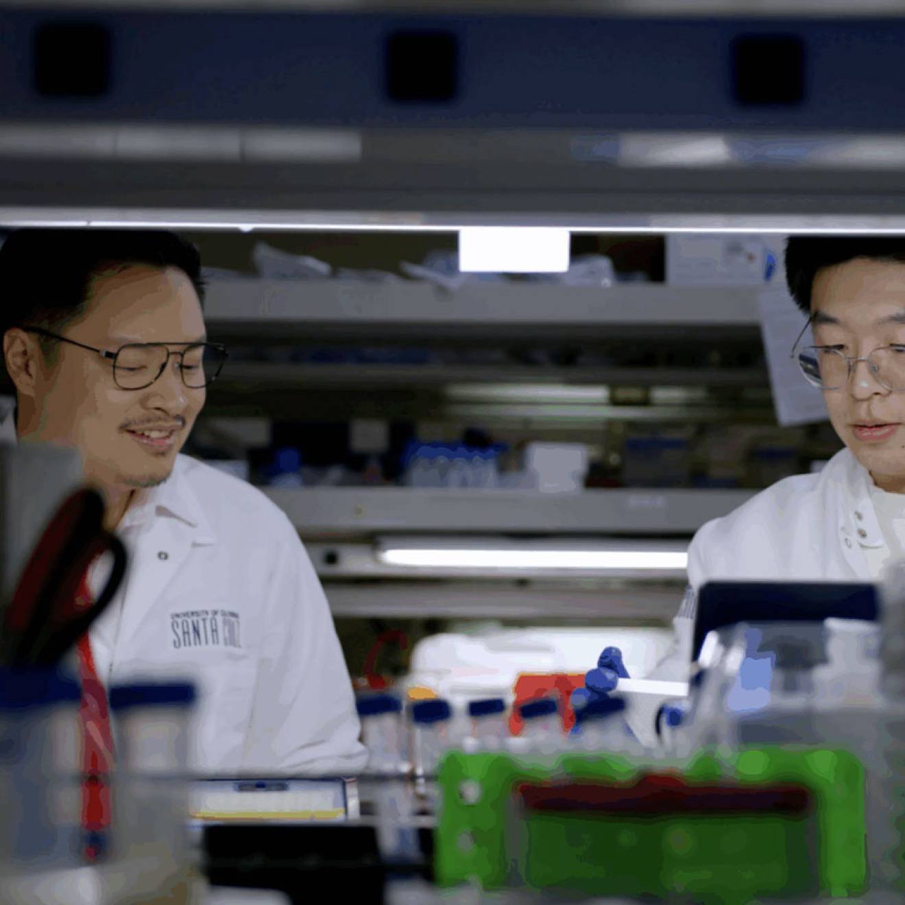 Two scientists in white coats work at a lab bench