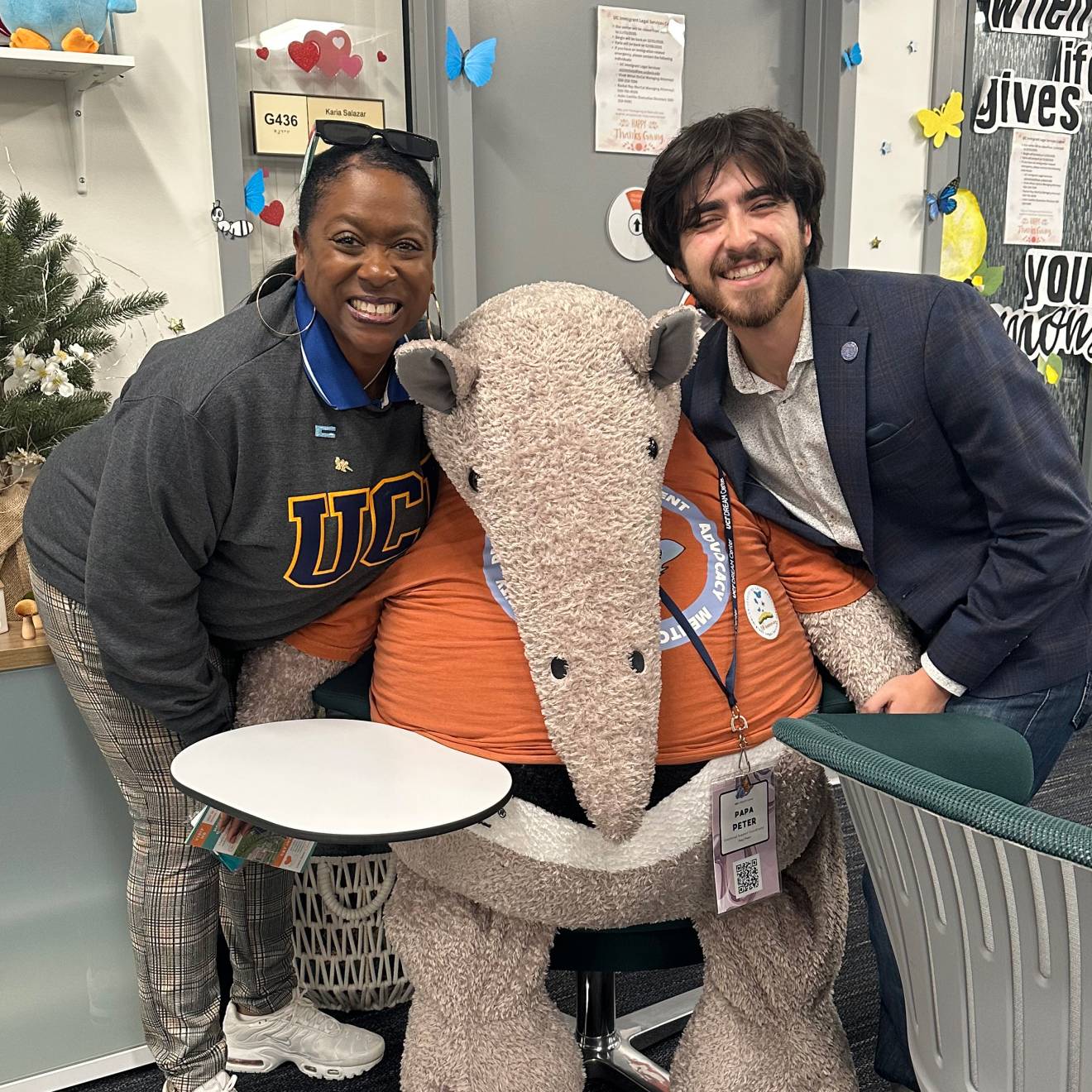 In a student center space, on the left, a woman with a sweatshirt that says UC and on the right, a young man with a beard smiling; center, a stuffed Anteater, the UC Irvine mascot