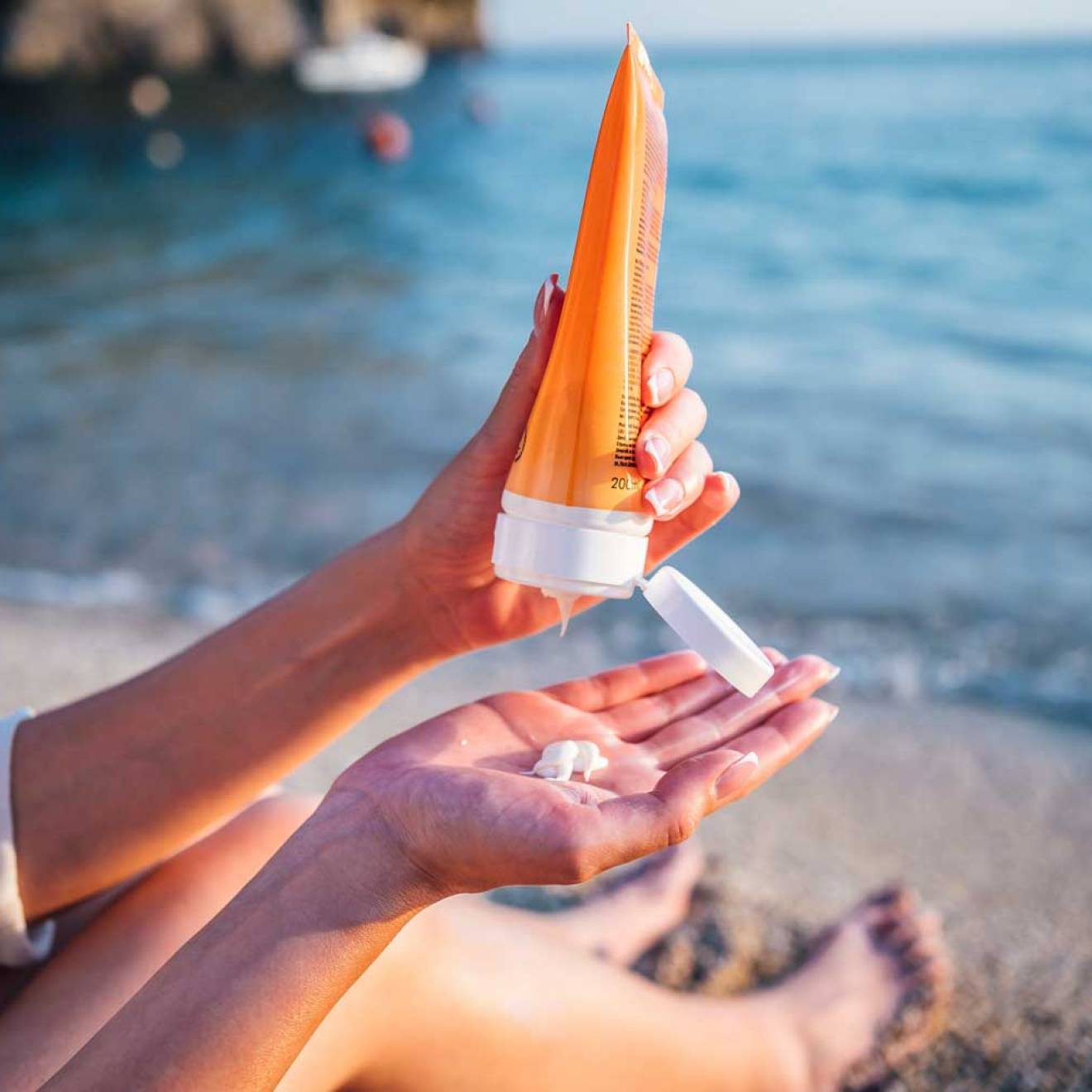 Someone sitting on the beach holds a tube of sunscreen in one hand with the other with a dab of sunscreen on it