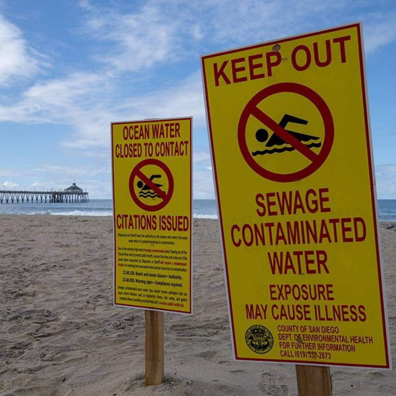 An empty beach with a pier in the background on a nice day bears signs that say Keep Out Sewage Contaminated Water and Ocean Water Closed to Contact Citations Issued