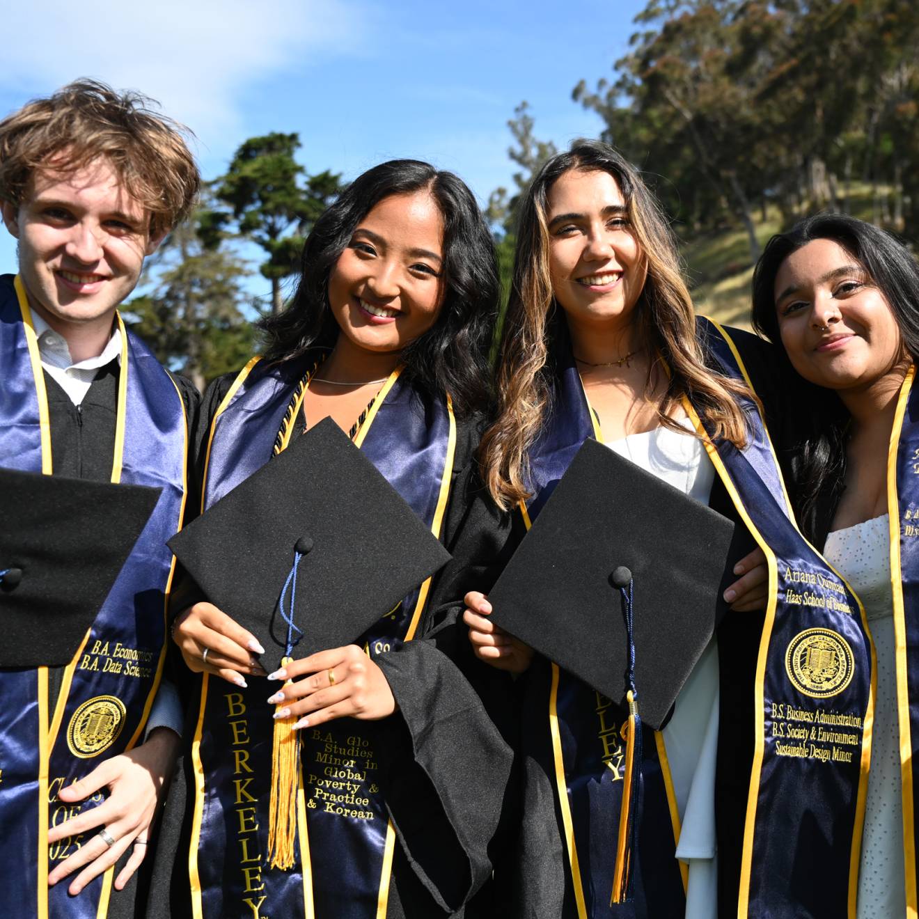 Four graduates in robes and stoles hold their caps and smile at the camera