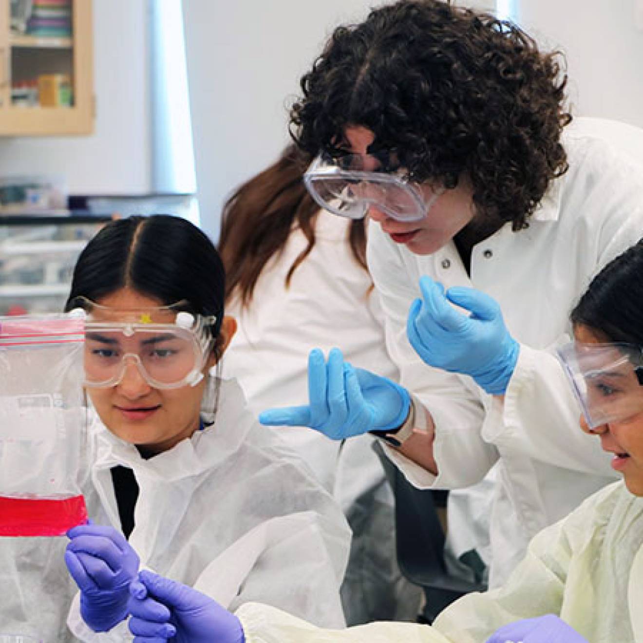 A college student in PPE shows two high schoolers in PPE how to collect a sample in a lab