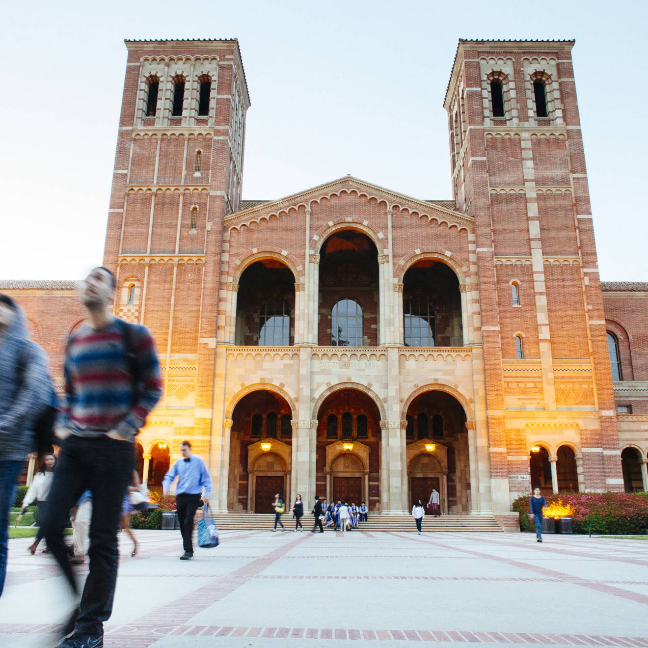 Students walk in front of a large building with two towers, Royce Hall, on the UC campus as evening begins to fall