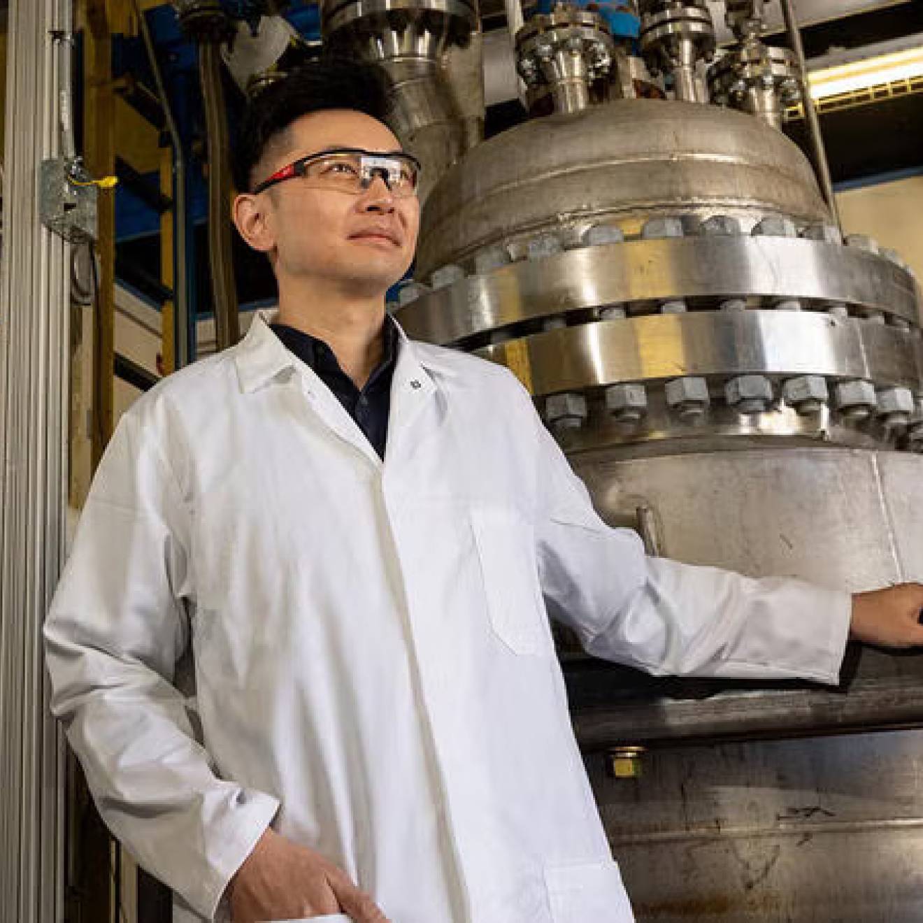 A professor in a white lab coat and safety goggles poses next to equipment
