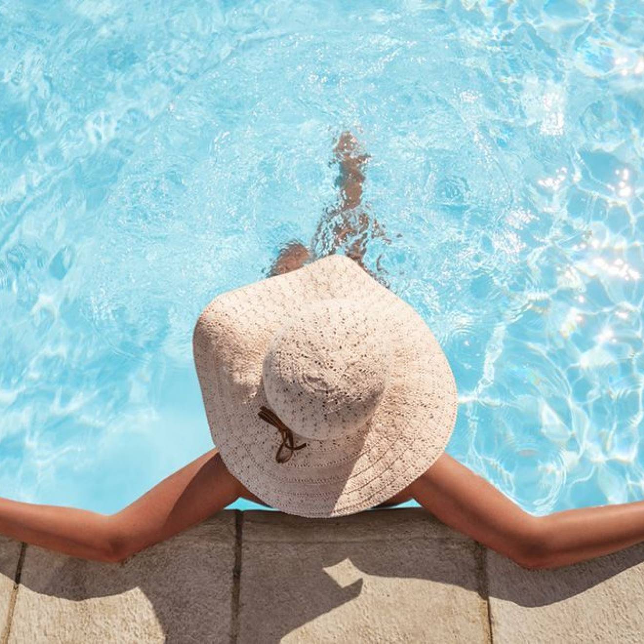 A woman wearing a brimmed straw hat relaxes at the edge of a swimming pool, photographed from above
