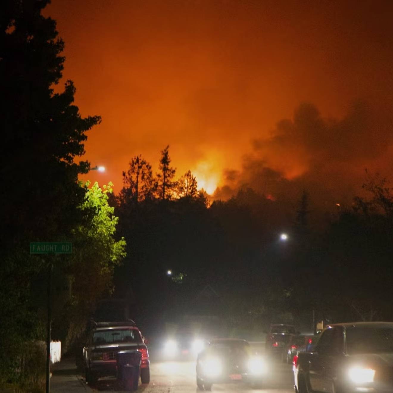 A line of cars, headlights on, sit in traffic as a red sky from a wildfire rages in the background
