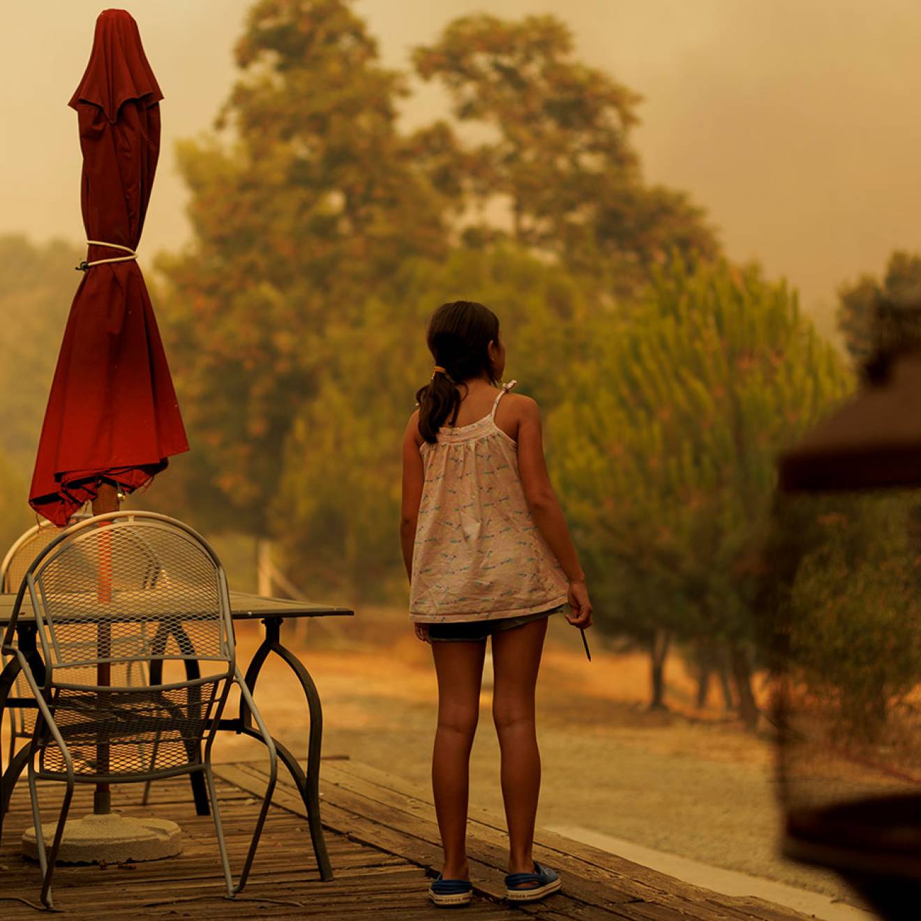 A school-aged girl photographed from behind, standing on the deck of her house, looking at a smoky sky